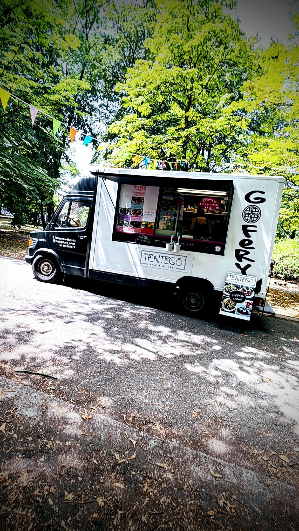 Główne zdjęcie Food Trucka TenTego - czarno-biały kultowy Mercedes w cieniu festiwalowych drzew z powiewającymi proporczykami i menu gofrów